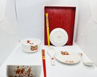 May include: A set of Chinese New Year themed dinnerware featuring a red and gold design with a butterfly motif. The set includes two bowls, two pairs of chopsticks, a spoon, a small plate, and a larger plate. The larger plate is in a red tray.