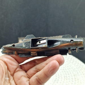 May include: A vintage pencil sharpener with a dark, weathered finish. The sharpener has a rectangular body with a small opening to insert a pencil. The metal has a mix of black and bronze tones, suggesting age and use.