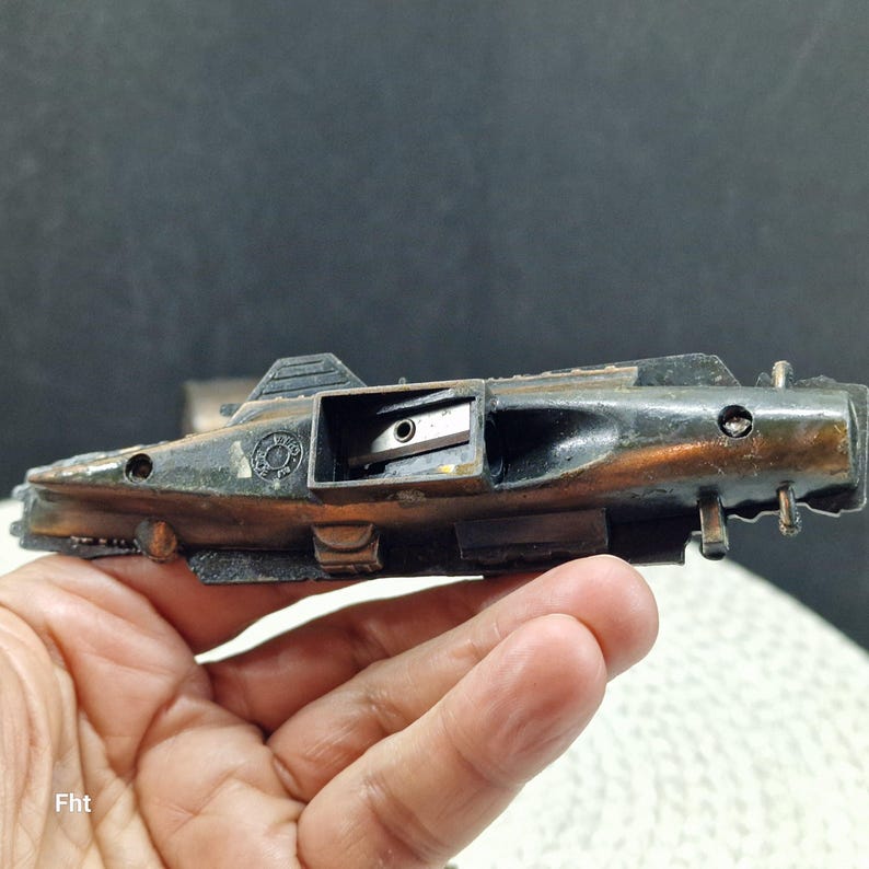 May include: A vintage pencil sharpener with a dark, weathered finish. The sharpener has a rectangular body with a small opening to insert a pencil. The metal has a mix of black and bronze tones, suggesting age and use.