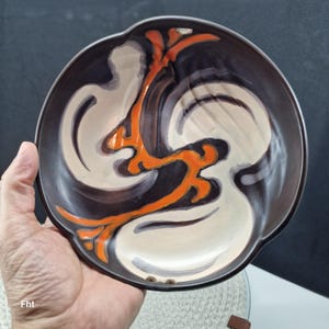 Op de afbeelding: Een keramische schaal met een donkerbruine rand en een centraal ontwerp met witte, zwarte en oranje abstracte vormen. De schaal heeft een enigszins onregelmatige vorm.