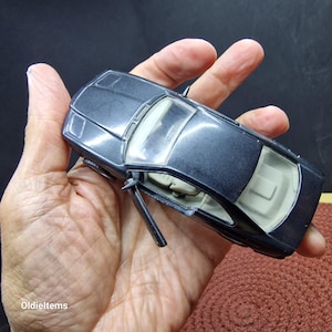 Puede incluir: Un coche de modelo fundido a presión de color gris oscuro con una puerta abierta, que revela un interior beige. El coche se sostiene en una mano, mostrando sus detalles. Es un objeto de colección vintage.