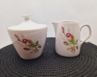 May include: A white sugar bowl and creamer set with a rose floral design. The set has a gold trim around the top of each piece.