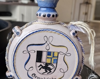May include: A white ceramic bottle with a blue and white lid and a white rope handle. The bottle has a blue and yellow crest with a black animal and the text "Brau bründer".