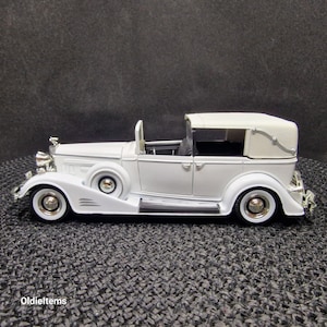 May include: White vintage car model with a white fabric roof and chrome accents. The car is a convertible with the top up. The car is on a black textured surface.