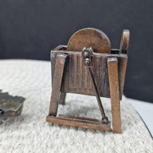 May include: A miniature bronze-colored pencil sharpener with a hand crank and a grinding wheel. The sharpener is rectangular with a small base and a handle. The wheel is visible at the top. The item is on a white textured surface.