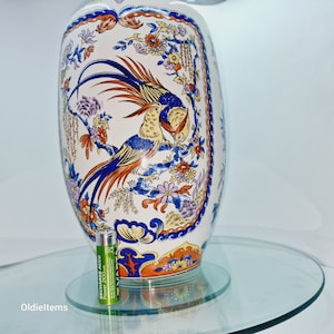 May include: A large white ceramic vase with a blue and orange floral design featuring two birds. The vase is decorated with a repeating pattern of flowers and leaves. The vase is sitting on a glass surface.
