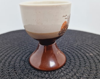 May include: A brown and white ceramic egg cup with a tree and bird design. The cup has a wide base and a narrow top.