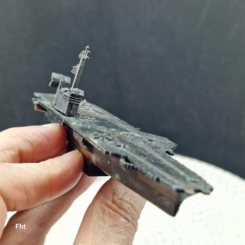 May include: A detailed model of a warship, primarily dark gray with visible weathering. The model features a detailed superstructure, including a mast and radar array. The ship's deck shows intricate detailing, suggesting a miniature replica of a naval vessel.