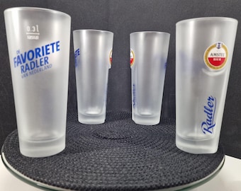 May include: Four frosted beer glasses with printed text and logos. One glass features "DE FAVORITE RADLER VAN NEDERLAND" in blue. Another has the Amstel Bier logo and the word "Radler" in blue. The other two glasses have the word "Amstel" in blue.