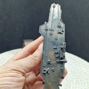 May include: A detailed, dark metal miniature of an aircraft carrier. The model features intricate details of the flight deck, including markings and miniature aircraft. The piece is held in a hand, showcasing its size and craftsmanship. The overall color is a dark, aged metal.