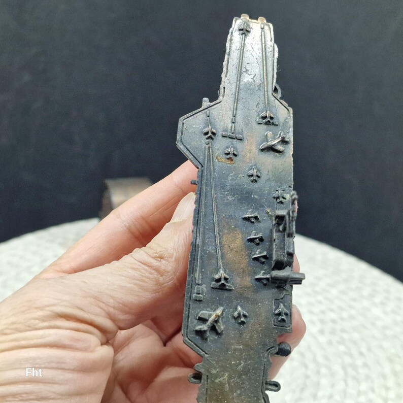 May include: A detailed, dark metal miniature of an aircraft carrier. The model features intricate details of the flight deck, including markings and miniature aircraft. The piece is held in a hand, showcasing its size and craftsmanship. The overall color is a dark, aged metal.