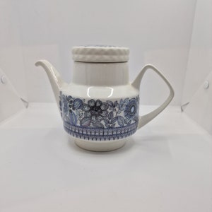 Könnte beinhalten: Eine weiße Teekanne aus Keramik mit einem blauen und violetten Blumenmuster. Die Teekanne hat einen Deckel und einen Ausguss.