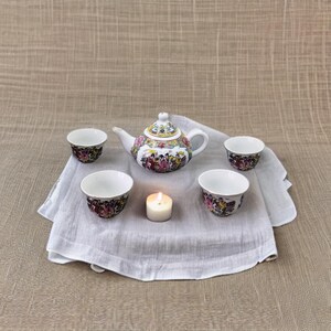 May include: A white porcelain teapot with a floral design and four matching cups. The teapot has a lid and a spout. The cups are small and have a handle. A white candle is in the center of the image.