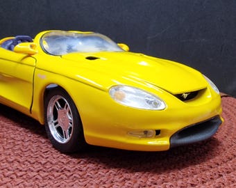 May include: A yellow die-cast model car of a Ford Mustang concept car. The car has a black interior and a black front bumper.
