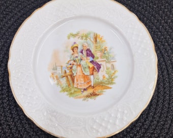 May include: A white porcelain plate with a gold rim features a hand-painted scene of a couple in a garden setting. The couple are dressed in 18th-century clothing and are surrounded by flowers and trees.