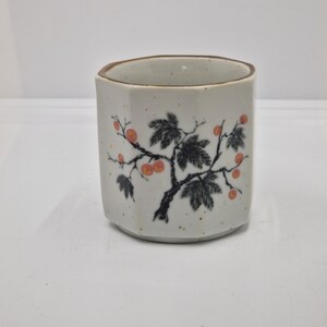 Könnte beinhalten: Weiße Keramiktasse mit braunem Rand und einem schwarzen und roten Blumendesign. Das Design zeigt einen Ast mit Blättern und roten Beeren.