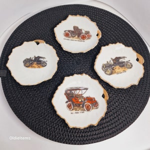 May include: Four white ceramic plates with gold trim, each featuring a vintage car illustration. The plates are on a black background. The cars are a 1908 Oldsmobile Curved Dash Runabout, a 1908 Ford Model T, a 1908 Ford Model T, and a 1908 Model T.