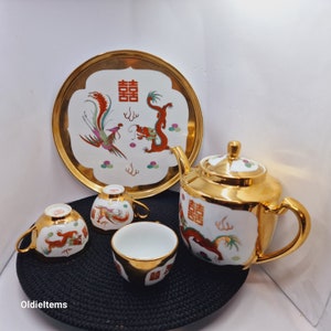 May include: A gold-trimmed white porcelain tea set with a dragon and phoenix design. The set includes a teapot, four cups, and a serving tray. The tray features the Chinese characters "囍" (double happiness) and "雙喜" (double joy).