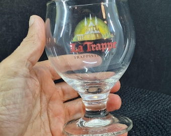 May include: A clear glass beer goblet with a flared rim and a short stem. The glass features the text "La Trappe" in red and a yellow and green graphic. The glass is held in a hand.
