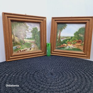Puede incluir: Dos pinturas de paisajes enmarcadas con marcos de madera. Las pinturas representan escenas rurales con árboles, campos y un río. Los marcos son de color marrón oscuro.
