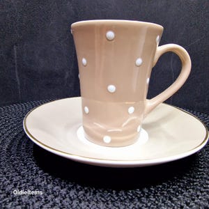 Könnte beinhalten: Eine beige Kaffeetasse mit weißen Punkten und einer passenden Untertasse. Die Untertasse hat einen goldenen Rand.