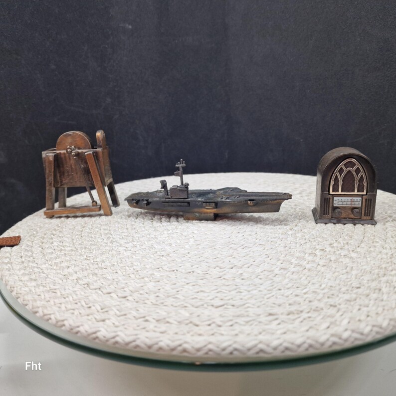 May include: Miniature metal sculptures of a ship, a radio, and a grinding wheel. The ship is dark gray with a small tower. The radio is brown with a decorative arched window. The grinding wheel is brown with a circular wheel.