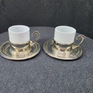 May include: Two white ceramic espresso cups with matching saucers, each encased in a polished silver-tone metal holder featuring a curved handle. The cups are cylindrical with a smooth finish, and the saucers have a simple, circular rim design.