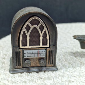 May include: A miniature antique-style radio, primarily bronze in color, with a detailed facade. The radio features a decorative arched window design and a dial with numbers. The radio's knobs and overall design evoke a vintage aesthetic.
