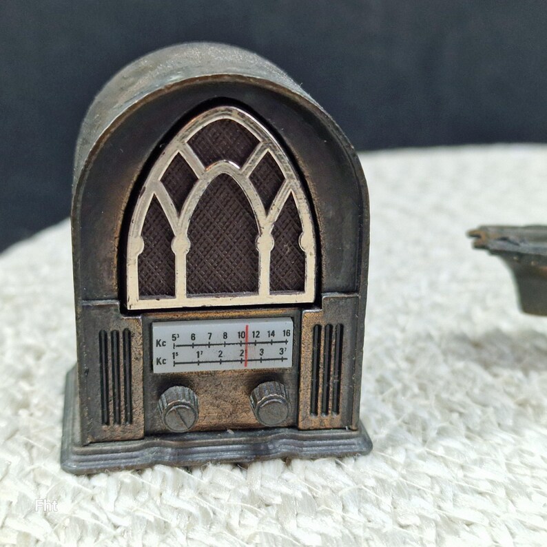 May include: A miniature antique-style radio, primarily bronze in color, with a detailed facade. The radio features a decorative arched window design and a dial with numbers. The radio's knobs and overall design evoke a vintage aesthetic.