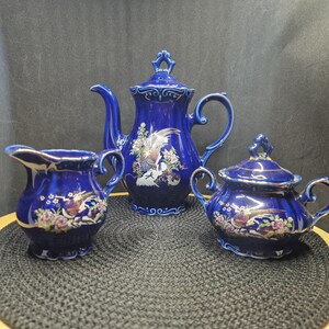 May include: A cobalt blue porcelain tea set featuring a teapot, creamer, and sugar bowl. Each piece is adorned with a gold trim and a detailed floral and bird design. The set is displayed on a black surface.