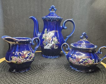 May include: A cobalt blue porcelain tea set featuring a teapot, creamer, and sugar bowl. Each piece is adorned with a gold trim and a detailed floral and bird design. The set is displayed on a black surface.