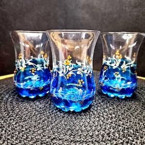 Könnte beinhalten: Drei klare Teetassen aus Glas mit blauem und weißem Blumendesign. Der Boden jeder Tasse ist mit einer leuchtenden blauen Farbe bemalt, mit weißen und goldenen floralen Akzenten. Die Tassen werden auf einer dunkelgrauen Oberfläche präsentiert.