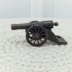 May include: A miniature bronze cannon with spoked wheels. The cannon barrel is detailed with ridges and a small ball at the end. The wheels and base are also bronze, creating a cohesive antique aesthetic. The cannon sits on a textured, off-white surface.