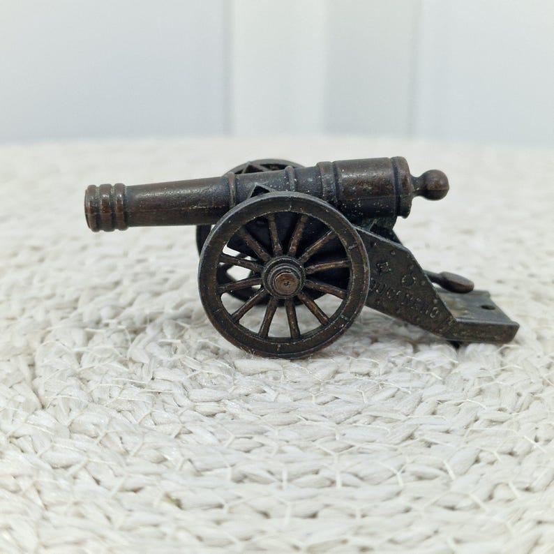 May include: A miniature bronze cannon with spoked wheels. The cannon barrel is detailed with ridges and a small ball at the end. The wheels and base are also bronze, creating a cohesive antique aesthetic. The cannon sits on a textured, off-white surface.