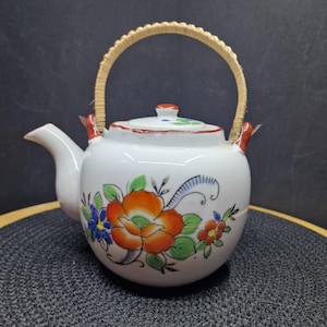Könnte beinhalten: Weiße Keramik-Teekanne mit geflochtenem Griff und floralem Design. Die Teekanne hat eine große orangefarbene Blume mit grünen Blättern, blauen und roten Blüten und einen roten Rand. Ausguss und Deckel sind weiß mit roten und grünen Akzenten.