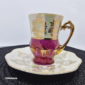 May include: A white and purple porcelain teacup and saucer set with gold floral accents. The cup has a burgundy base and a white top with gold floral designs. The saucer has a white base with gold floral designs.