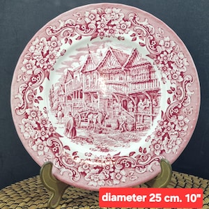 Puede incluir: Plato decorativo de 25 cm de diámetro. Borde floral rojo y blanco y escena central de un pueblo histórico con edificios y figuras. El plato se exhibe sobre un soporte de latón.