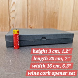 Op de afbeelding: Een donkergrijze wijnkurkopener set. De set zit in een rechthoekige doos van 20 cm lang, 16 cm breed en 3 cm hoog. Een rode en gele Kodak-batterij staat naast de doos. De tekst "wine cork opener set" is zichtbaar.