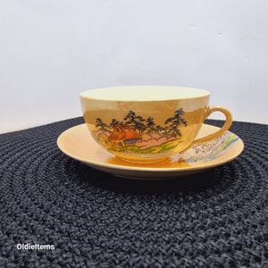 Może przedstawiać: Zestaw vintage filiżanki i spodeczka z złotym i białym tłem oraz malowanym obrazem domu i drzew. Filiżanka i spodeczek mają delikatny wzór kwiatowy.