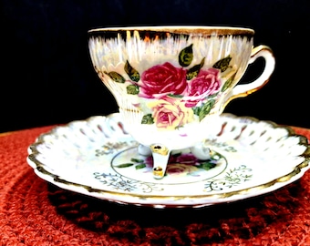 Puede incluir: Un juego de taza y platillo de té vintage. La taza presenta un diseño floral con rosas rojas y amarillas, ribete dorado y un asa delicada. El platillo tiene un patrón floral a juego y ribete dorado. El juego se exhibe sobre una superficie roja.