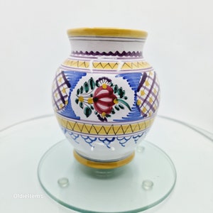 Puede incluir: Un pequeño jarrón de cerámica blanca con cuerpo redondo y borde amarillo. El jarrón está decorado con un patrón de cuadros azules y amarillos, un diseño floral en tonos rojos, verdes y amarillos, y un patrón festoneado amarillo y azul.