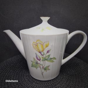 Könnte beinhalten: Weiße Porzellan-Teekanne mit gelbem Blumendesign. Die Teekanne hat einen Deckel und einen Henkel.