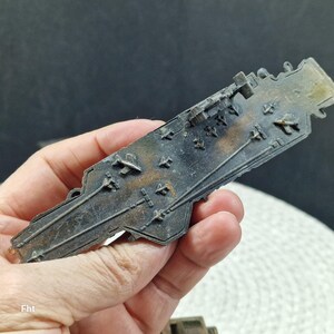 May include: A detailed, miniature metal aircraft carrier model with a weathered, aged appearance. The model features intricate details of the flight deck, including tiny aircraft and deck markings. The color is a mix of dark gray and brown.