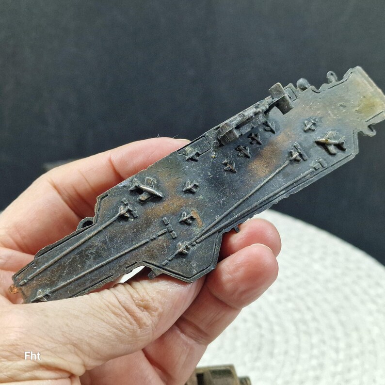 May include: A detailed, miniature metal aircraft carrier model with a weathered, aged appearance. The model features intricate details of the flight deck, including tiny aircraft and deck markings. The color is a mix of dark gray and brown.