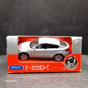 Può includere: Un'auto modello in metallo pressofuso in scala 1:43 di colore argento di una BMW X6 nella sua confezione originale. La scatola è rossa e bianca con le parole "Welly Nex Model" e "Die Cast Metal Scale Model" stampate su di essa.