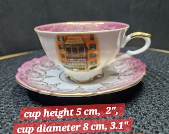 May include: A delicate teacup and saucer set. The white cup, 5 cm tall, displays an architectural scene and gold detailing. The pink-edged saucer, 12 cm in diameter, matches the cup's design. The cup's diameter is 8 cm.