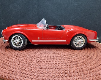 May include: A red toy car model of a classic convertible sports car. The car has a black interior and chrome wheels. The car is sitting on a red braided rug.