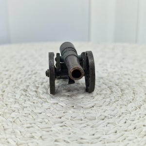 May include: A miniature bronze cannon with two wheels. The cannon barrel is centered, and the wheels are on either side. The cannon is set against a textured, off-white surface.