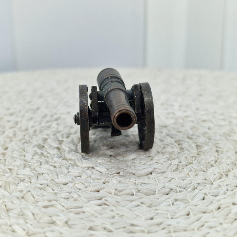 May include: A miniature bronze cannon with two wheels. The cannon barrel is centered, and the wheels are on either side. The cannon is set against a textured, off-white surface.