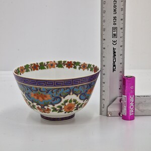 May include: A white porcelain bowl with a blue and gold floral pattern. The bowl is approximately 7.5 cm in diameter and is sitting on a white surface next to a ruler and a pink battery.